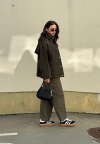 Ελαιόχρωμο αδιάβροχο μπουφάν με κουμπιά αρμάτωσης, συνδυασμένο με ελαιόχρωμες παντελόνες. Μαύρη τσάντα ώμου και μαύρα αθλητικά παπούτσια με λευκές ρίγες.