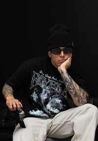 Černé oversized grafické tričko s potiskem "FAVELA", světlé béžové cargo kalhoty, černá čepice, sluneční brýle a fotoaparát v ruce.