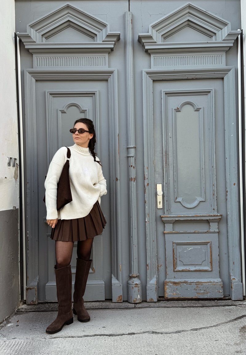 Witte geribbelde trui, bruine plisserok tot mini-lengte, donkerbruine knieën-hoge laarzen. Achtergrond toont een gestructureerde grijze dubbele deur met detaillering.