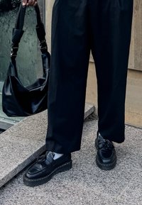 Sapatos de couro pretos com sola grossa e detalhes franjados, combinados com calças largas pretas e uma bolsa preta. Superfície texturizada visível.
