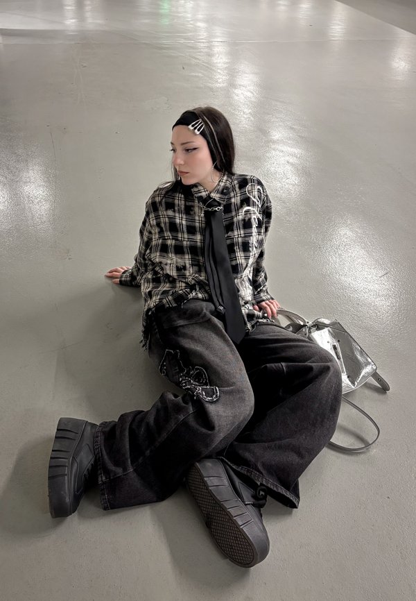Nooriku juustega ja juuksenõeltega noor istub sileda põranda peal, kandes musta-valget ruudulist särki, musta lipsu, laiade säärega pükse ja platvormjalatseid.