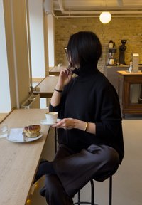 Pull noir en maille avec un col montant, associé à un pantalon marron ample. Tient une tasse de café sur une soucoupe blanche, à côté d'une pâtisserie sur une assiette.