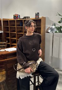 Bruine gebreide trui met oversized mouwen, gecombineerd met een crèmekleurige kanten rand en zwarte broek met wijde pijpen. Houdt een beige handtas met patroon vast.