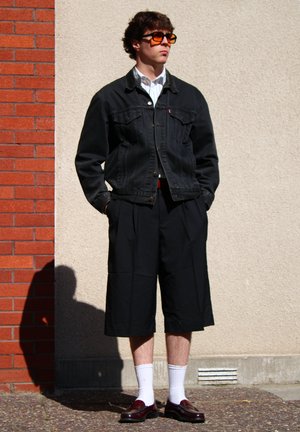 Chaqueta de mezclilla negra, camiseta a rayas, pantalones cortos negros con pliegues, calcetines blancos y zapatos oscuros con detalles decorativos, colocados sobre un fondo texturizado.