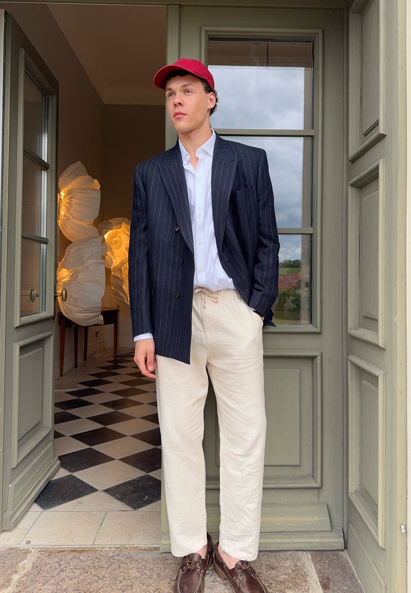 Young man in red cap wears navy pinstripe blazer, white shirt, cream trousers, and brown shoes, standing in a doorway with patterned floor inside.