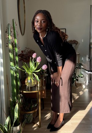 Mujer con blusa negra y falda marrón se inclina hacia adelante cerca de una pequeña mesa con tulipanes rosas y plantas altas verdes en una habitación iluminada por el sol.