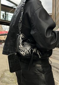 Bolso de mano de cuero negro con un acabado texturizado, que cuenta con cremalleras, tachuelas y una correa. Incluye una pequeña billetera adjunta en material a juego.