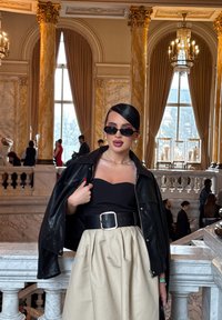 Μαύρο δερμάτινο τζάκετ πάνω από ένα μαύρο strapless τοπ, φιξαρισμένο με μια φαρδιά ζώνη, συνδυασμένο με μια μπεζ πλισέ φούστα. Γυαλιά ηλίου και λείο μαλλί ολοκληρώνουν την εμφάνιση.