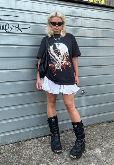 Camiseta gráfica negra con un estampado detallado de una mujer a caballo, combinada con pantalones cortos blancos con volantes y botas negras hasta la rodilla.