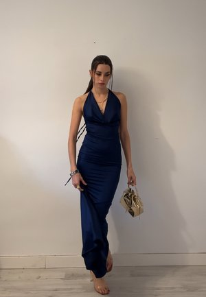 Mujer con un vestido halter azul marino que sostiene un pequeño bolso dorado, de pie descalza contra una pared blanca lisa.