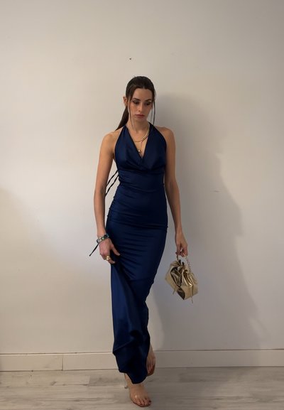 Mujer con un vestido halter azul marino que sostiene un pequeño bolso dorado, de pie descalza contra una pared blanca lisa.