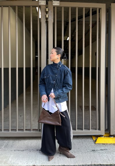 Mujer con chaqueta vaquera, camiseta blanca y pantalones anchos negros sosteniendo un bolso marrón, de pie frente a una puerta metálica cerrada en un entorno urbano.