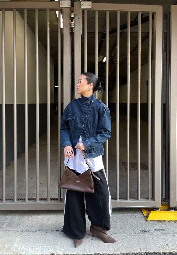 Vrouw in spijkerjack, wit shirt en zwarte wijde broek, die een bruine handtas vasthoudt, staande voor een gesloten metalen poort in een stedelijke omgeving.