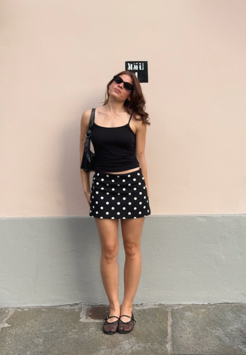 Jeune femme portant un débardeur noir, une jupe à pois, des lunettes de soleil noires et un sac d'épaule, debout contre un mur pâle avec un petit panneau au-dessus de sa tête.