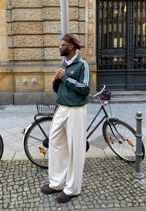 Chaqueta atlética verde con rayas blancas, pantalones anchos blancos, boina marrón y zapatos oscuros. Una bicicleta está en el fondo.
