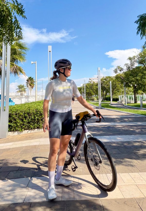 Un cycliste portant un casque se tient à côté d’un vélo de route sur une promenade ensoleillée bordée d’arbres.