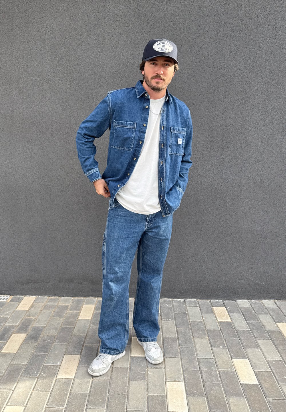 Jeanshemd mit Knopfverschluss, langen Ärmeln und zwei Brusttaschen; kombiniert mit locker sitzenden Jeans. Graue Turnschuhe und eine dunkelblaue Kappe runden das Outfit ab.