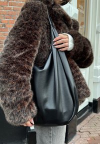 Mustat nahkainen hobo-laukku, jossa on pehmeä rakenne, suuri aukko ja vähäinen metalliosuus, yhdistettynä ruskeaan turkistakkiin ja vaaleanharmaisiin pantsereihin.