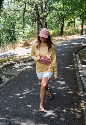 Camiseta amarilla de manga larga, pantalones cortos blancos, riñonera rosa y gorra. Sendero rodeado de árboles, con luz solar moteada en el suelo.