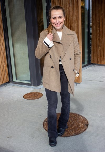 Abrigo marrón de doble botonadura con solapas, botones negros, usado sobre un suéter blanco. Combinado con pantalones de campana negros y zapatos negros.