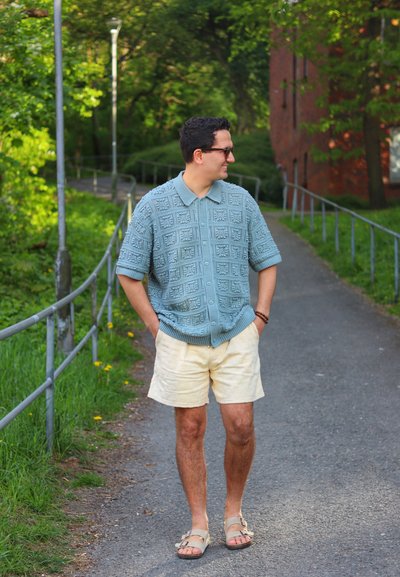 Suéter corto de manga corta de color azul claro con textura y cuello, shorts crema y sandalias marrones. Camino con vegetación y un edificio al fondo.