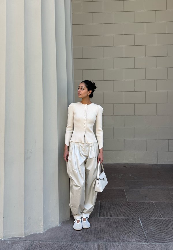 Femme en haut blanc à manches longues, pantalon ample de couleur claire et chaussures blanches tenant un sac à main blanc contre un mur à colonnes.