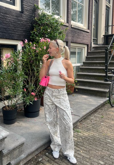 Top corto blanco de canalé, pantalones anchos de estampado de serpiente, bolso rosa y zapatillas blancas. Fondo de flores y ladrillo oscuro.