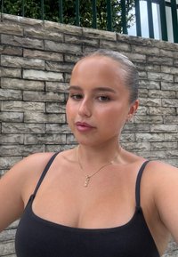 Musta hihaton paita. Kultainen käärmekoru kaulassa, lyhyet hiukset taaksepäin kammattuna. Vaalea ihonsävy ja taustalla teksturoitu kiviseinä.