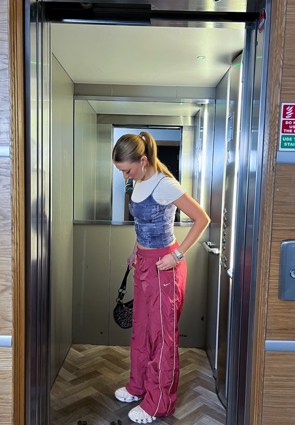 Tânără cu coadă de cal, în adidași albi, pantaloni roșii și bluză albastră, privind în jos într-un lift metalic.