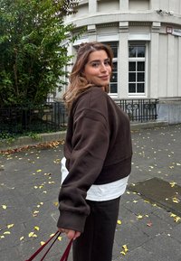 Καφέ κοντό sweatshirt με μακριά μανίκια, τοποθετημένο πάνω από μια λευκή μπλούζα, που συνδυάζεται με σκούρα παντελόνια και μια μπορντό τσάντα, μπροστά σε αστικό φόντο.