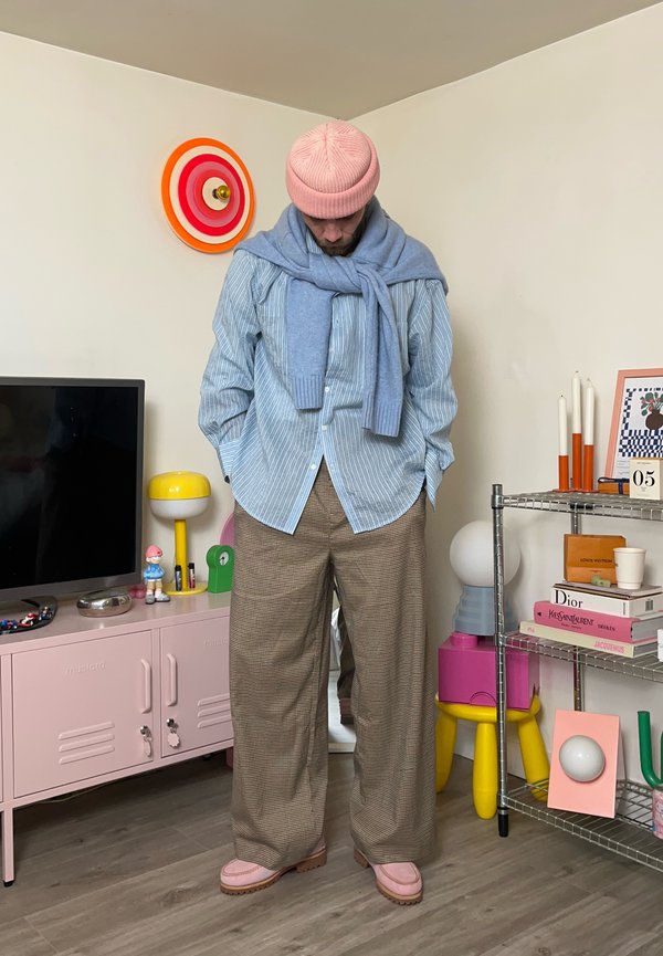 Man draagt een roze muts, blauw gestreept shirt, grijze trui om de schouders en bruine broek met wijde pijpen, staat in een kleurrijke, versierde kamer.