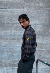 Μαύρο μπουφάν με διαμαντένιο μοτίβο, κίτρινες λεπτομέρειες και λογότυπο στο μανίκι. Φορεμένο πάνω από σκούρες παντελόνες, μπροστά σε μια υφασμάτινη γκρι τοίχο.