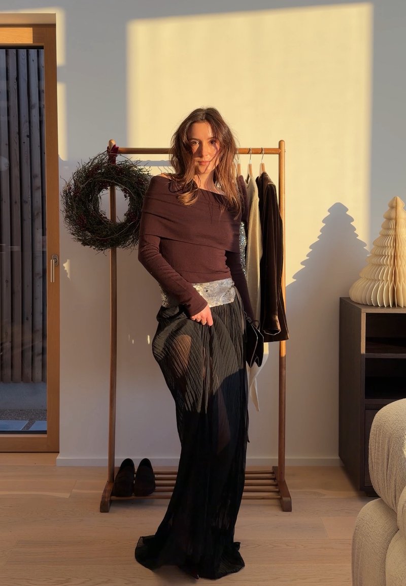 Femme debout à l'intérieur, portant une jupe noire transparente et un haut à manches longues, près d'un porte-vêtements en bois avec des vêtements suspendus et une couronne décorative.