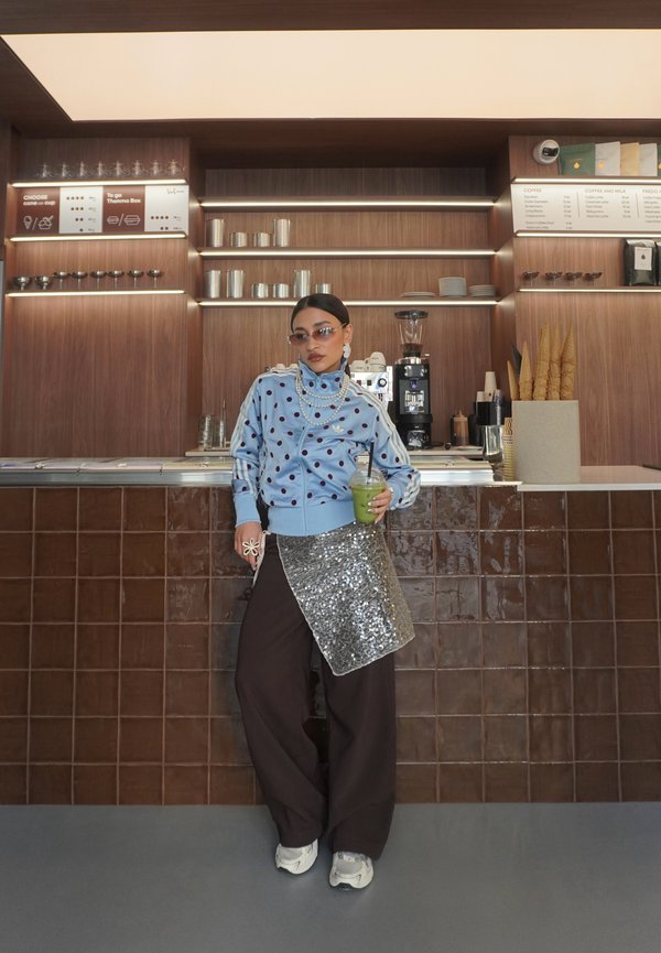 Femme portant une veste bleue à pois et une jupe à sequins, debout au comptoir du café, tenant une tasse de smoothie vert.