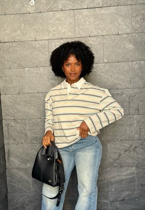 Mujer con cabello rizado, camiseta de manga larga a rayas y jeans claros, de pie contra una pared de piedra gris, sosteniendo un bolso negro con una mano.