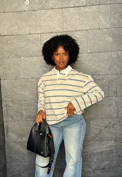 Mujer con cabello rizado, camiseta de manga larga a rayas y jeans claros, de pie contra una pared de piedra gris, sosteniendo un bolso negro con una mano.