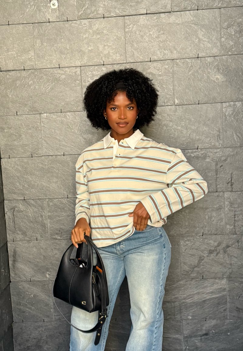 Frau mit lockigem Haar, die ein gestreiftes Langarmshirt und helle Jeans trägt, steht vor einer grauen Steinmauer und hält eine schwarze Handtasche in einer Hand.