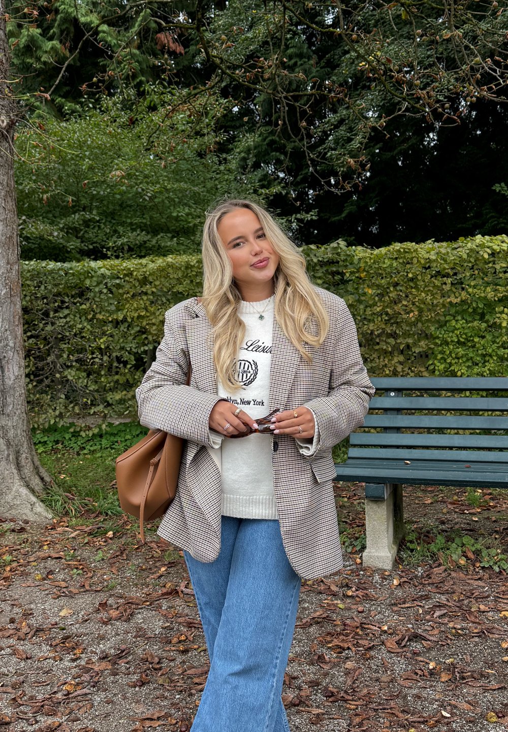 Ruudullinen bleiseri ruskean ja valkoisen sävyissä valkoisen collegepaidn päällä, yhdistettynä sinisiin levenevä lahkeisiin farkkuihin. Kädessä ruskea käsilaukku ja aurinkolasit.