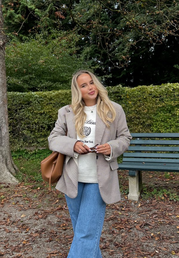 Rutete blazer i brune og hvite nyanser over en hvit genser, kombinert med blå vide jeans. Brun håndveske og solbriller i hånden.