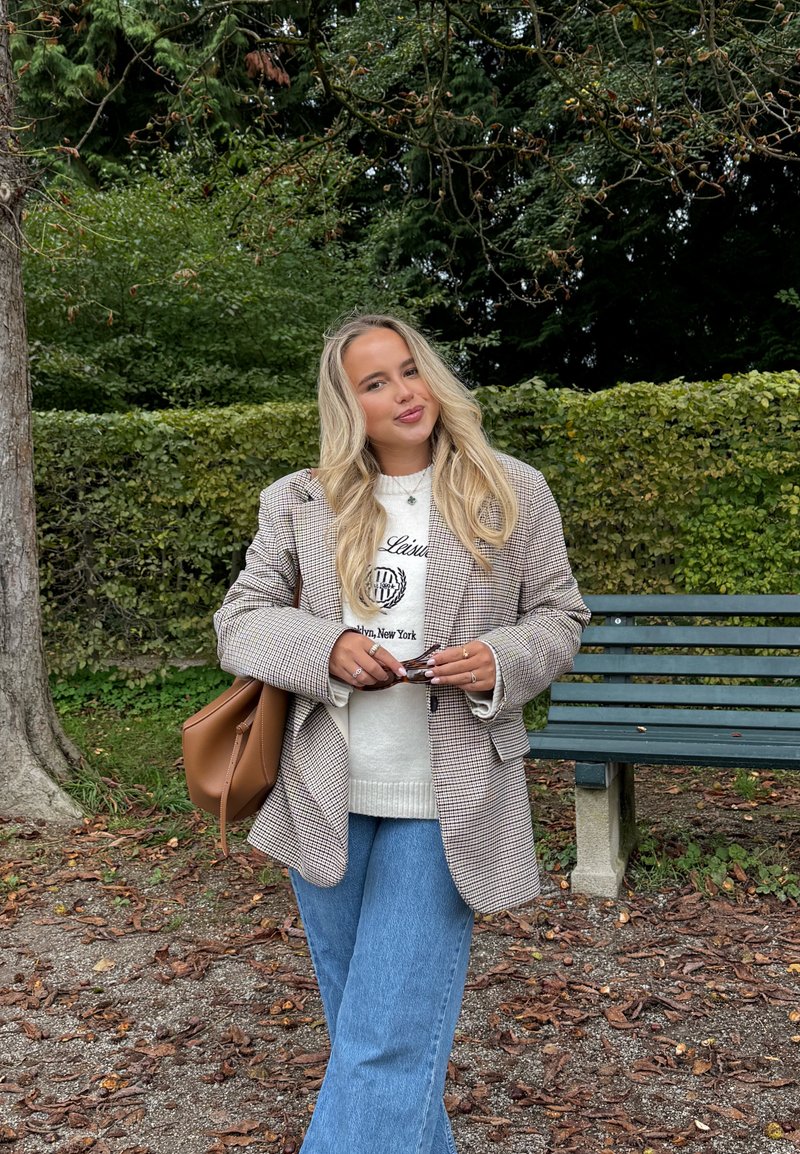 Karierter Blazer in Braun- und Weißtönen über einem weißen Sweatshirt, kombiniert mit blauen Schlagjeans. Braune Handtasche und Sonnenbrille in der Hand.