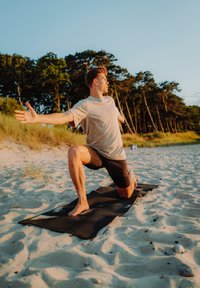 Muškarac u svijetlosivoj majici i crnim kratkim hlačama izvodi jogu na crnom prostircu na pješčanoj plaži sa drvećem u pozadini.