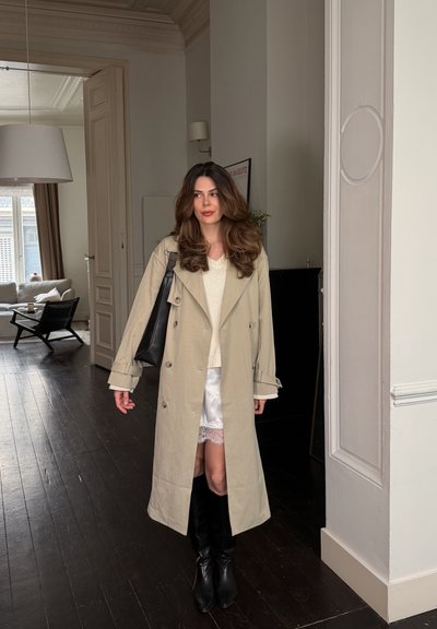 Mujer con cabello largo y castaño, vistiendo un abrigo beige, vestido blanco, botas negras y llevando un bolso negro de hombro en una sala de estar moderna.