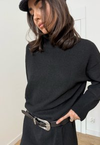 Pull noir en maille à coupe décontractée, porté par-dessus une ceinture marron avec une boucle argentée décorative, sur un fond clair.