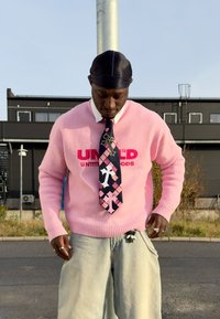 Homme portant un pull rose avec un texte rouge, une cravate à motifs de croix, un jean clair et un durag noir, debout à l'extérieur près d'un poteau et de bâtiments industriels.