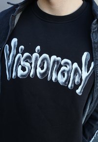 Hanorac negru din bumbac, prezentând litere albe îndrăznețe, ondulate, care spun "Visionary". Materialul pare moale, cu o textură fină.