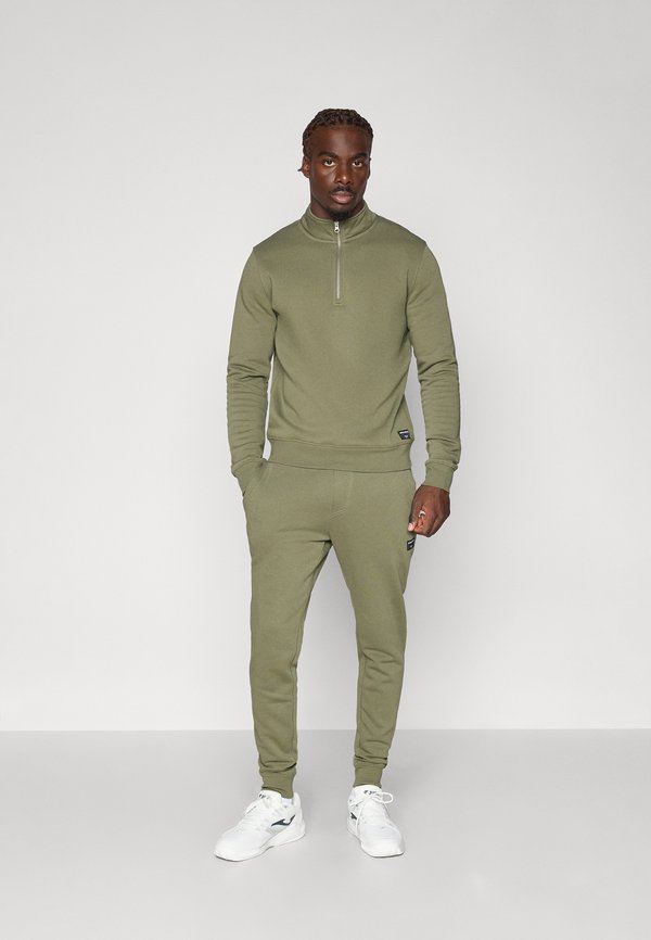 Chándal verde oliva con cuello con cremallera y puños acanalados. Los pantalones tienen un corte ajustado y bolsillos laterales. Se lleva con zapatillas blancas.