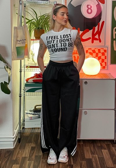 Camiseta corta blanca con texto en negrita negro, combinada con pantalones deportivos negros que tienen rayas blancas a los lados. Zapatillas blancas y una bolsa tote son visibles.