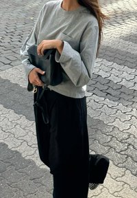 Mulher com uma camisa de manga longa cinza e calças pretas segurando uma carteira de mão preta, em pé sobre um pavimento com padrão.