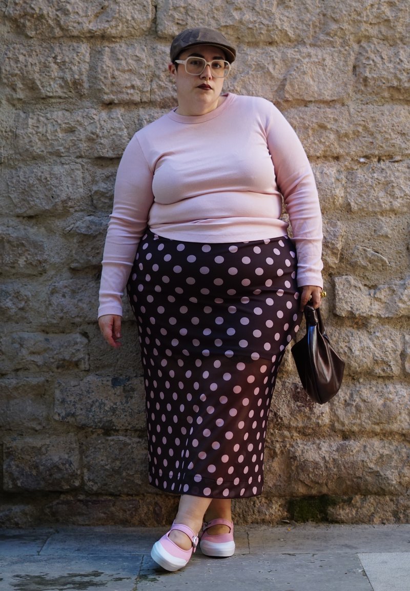 Personne portant un haut à manches longues rose pâle, une jupe noire à pois roses, des chaussures roses, une casquette brune et tenant un sac brun foncé près d'un mur en pierre.