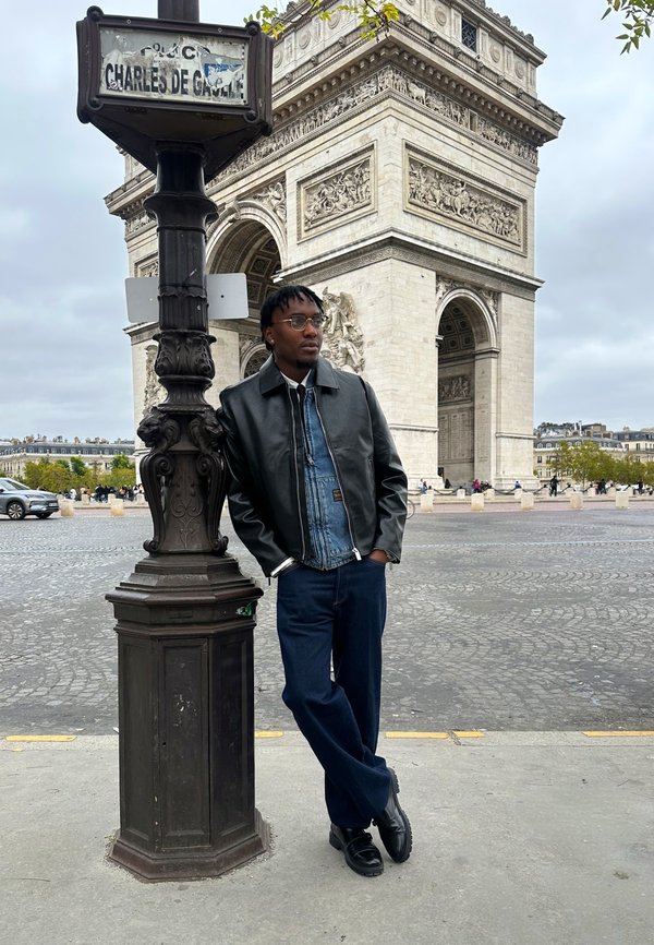 Un homme portant une veste en cuir noire pose à côté d’un panneau de rue devant l’Arc de triomphe à Paris.